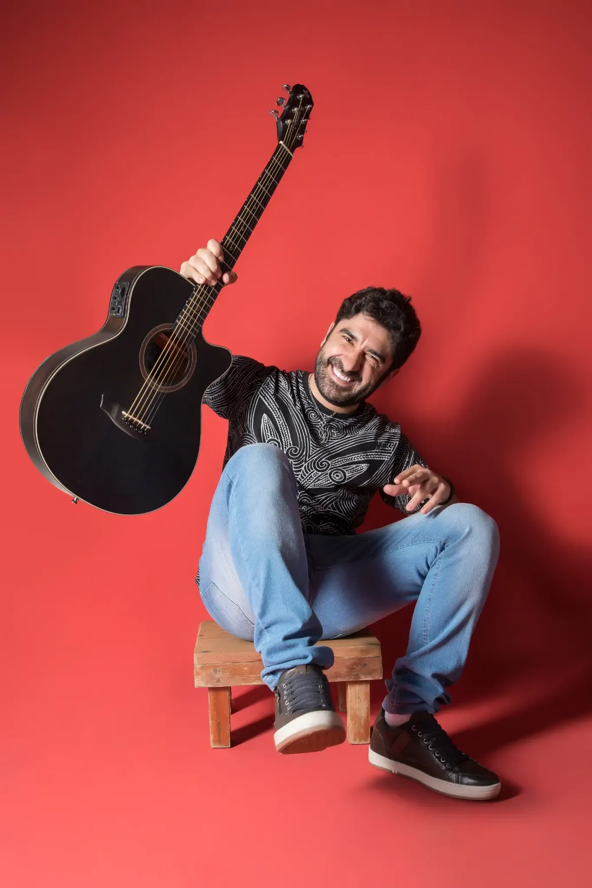 Ramonzera sorrindo, com seu violão, em ensaio de fundo vermelho
